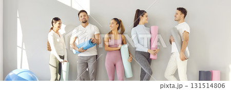 Group of young people in sportswear with exercise mats standing by gym wall and talking Group of young people in sportswear with exercise mats standing by gym wall and talking 131514806