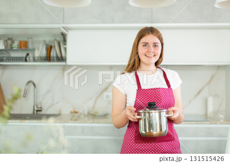 Happy teen girl wearing red polka dot apron and holding small pot after cooking in modern kitchen Happy teen girl wearing red polka dot apron and holding small pot after cooking in modern kitchen 131515426