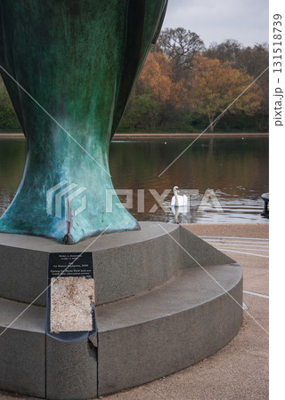 A green patinated sculpture on a stone pedestal with a plaque near a calm body of water, a white swan, and autumn colored trees in a London park. A green patinated sculpture on a stone pedestal with a plaque near a calm body of water, a white swan, and autumn colored trees in a London park. 131518739