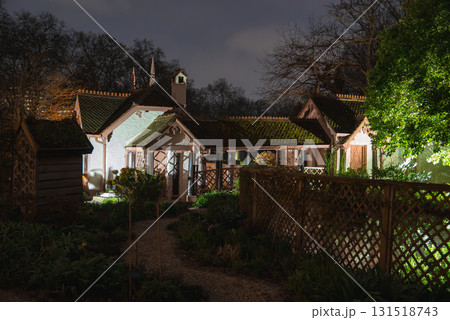 A historic cottage in London illuminated at night, featuring steep roofs, decorative trim, a chimney, and a lush garden with wooden lattice fencing. 131518743