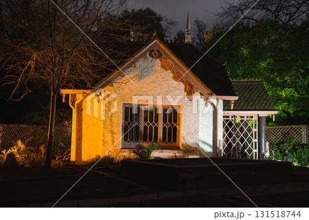 A quaint cottage with a white facade, wooden trim, and steep roof, surrounded by garden beds and trees, illuminated against a nighttime backdrop. A quaint cottage with a white facade, wooden trim, and steep roof, surrounded by garden beds and trees, illuminated against a nighttime backdrop. 131518744