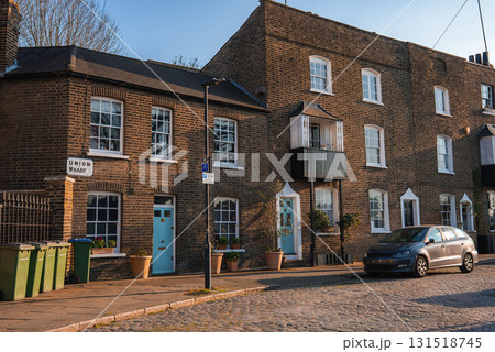 Row of brick townhouses with colorful doors, cobblestone street, potted plants, parked car, and bare trees in late autumn or winter. 131518745
