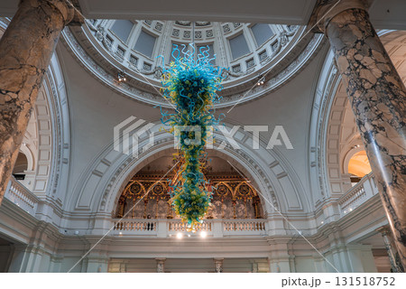 The grand interior of the Victoria and Albert Museum in London features a vibrant glass chandelier by Dale Chihuly, ornate arches, and marble columns. 131518752