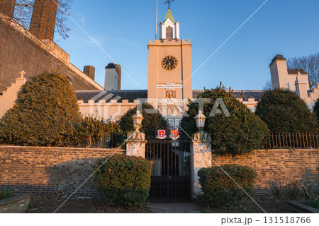 The Royal Hospital Chelsea in London features a central clock tower, golden clock face, ornate iron gate, trimmed hedges, and a cream facade under blue skies. 131518766