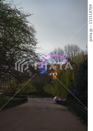 A tranquil park pathway in London bordered by trees and greenery, with the London Eye glowing in purple and blue against a dim evening sky. A tranquil park pathway in London bordered by trees and greenery, with the London Eye glowing in purple and blue against a dim evening sky. 131518769