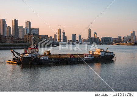 View of the River Thames at sunset in London, featuring anchored boats, a barge, and the modern skyscrapers of Canary Wharf reflecting warm light. View of the River Thames at sunset in London, featuring anchored boats, a barge, and the modern skyscrapers of Canary Wharf reflecting warm light. 131518780