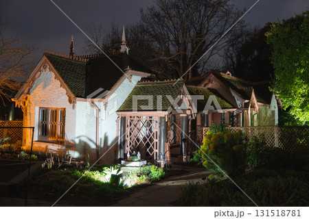 A cottage style building with pointed gables and decorative woodwork, surrounded by a garden with lattice fencing, set against bare trees at night. A cottage style building with pointed gables and decorative woodwork, surrounded by a garden with lattice fencing, set against bare trees at night. 131518781