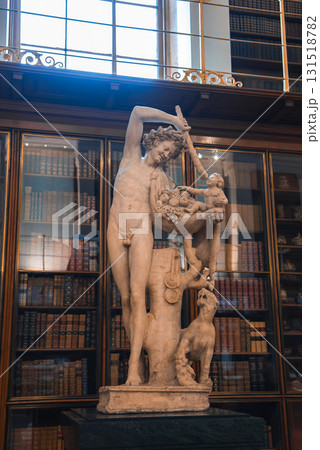 A marble statue of a male figure holding a fruit basket, with a child and dog at his feet, displayed indoors among tall wooden bookshelves and large windows. A marble statue of a male figure holding a fruit basket, with a child and dog at his feet, displayed indoors among tall wooden bookshelves and large windows. 131518782