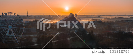 A warm sunrise illuminates Riga, Latvia, featuring the National Library, St. Peter's Church spire, a Ferris wheel, and the misty Daugava River. 131518793