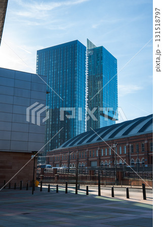 View of Manchester featuring Deansgate Square towers, a historic red brick building, a paved area with bollards, and a clear blue sky. 131518797