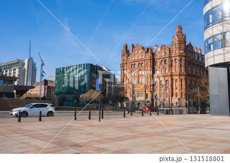 View of Manchester featuring the historic Midland Hotel, a modern glass building, a clean plaza with cars and pedestrians, and a clear blue sky. 131518801