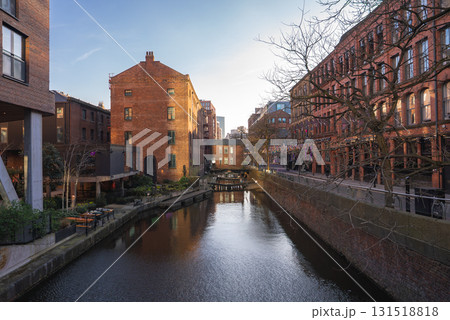 A tranquil canal in Manchester, England, bordered by historic red brick buildings, tree lined pathways, and outdoor seating under warm sunset lighting. 131518818