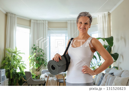 Beautiful gray-haired middle-aged mature woman holds a rolled yoga mat at home. Beautiful gray-haired middle-aged mature woman holds a rolled yoga mat at home. 131520823