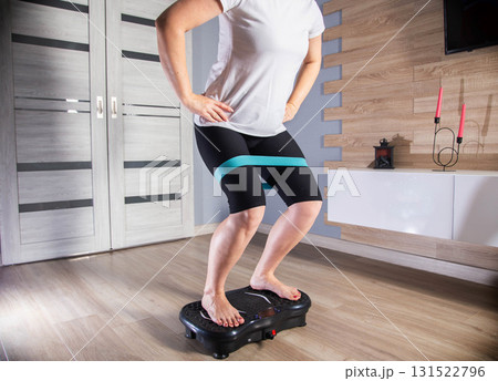 Brunette girl exercises on a modern vibration platform with a rubber band for fitness at home. Concept of fitness and sports on a vibration platform, aerobics Brunette girl exercises on a modern vibration platform with a rubber band for fitness at home. Concept of fitness and sports on a vibration platform, aerobics 131522796
