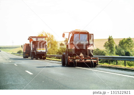A tractor with a brush sweeps the road against the backdrop of a milling machine cutting asphalt. Country road repairs. Copy space for text, industry A tractor with a brush sweeps the road against the backdrop of a milling machine cutting asphalt. Country road repairs. Copy space for text, industry 131522921