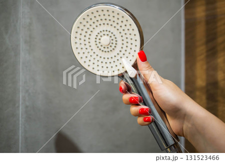 female hand with red manicure holding shower head in hand against grey tile background in bathroom, close-up 131523466