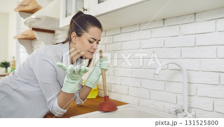 Young brunette woman cleaning blocked sink and drain with a plunger in kitchen at home. Young brunette woman cleaning blocked sink and drain with a plunger in kitchen at home. 131525800