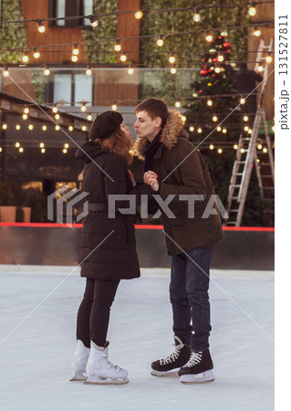 A young couple in love kissing on an ice rink against the background of retro garlands with light bulbs and a Christmas tree in winter 131527811