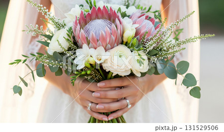 A hyper-realistic, high-detail close-up photograph of a bride's hands gently holding a gorgeous wedding bouquet filled with pale pink protea 'Blushing Bride', ivory Freesias, soft foliage A hyper-realistic, high-detail close-up photograph of a bride's hands gently holding a gorgeous wedding bouquet filled with pale pink protea 'Blushing Bride', ivory Freesias, soft foliage 131529506