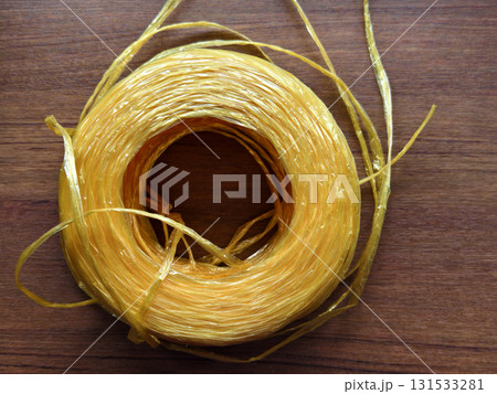 Close-up of yellow raffia plastic rope roll on wooden surface. Strong, durable, and flexible cord used for packaging, shipping, agriculture, DIY, and craft projects. 131533281