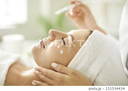 Beautician's hands applying anti-aging facial cream on woman client face in spa salon. 131534434