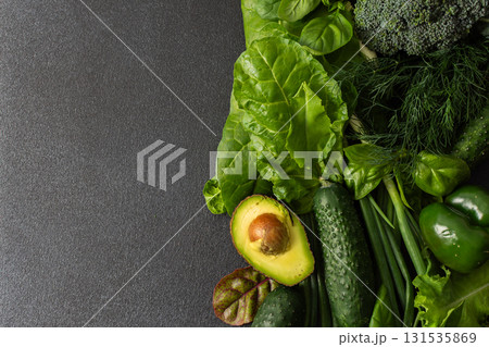 Displaying a vibrant assortment of fresh greens and vegetables on a dark surface, this scene features cucumbers, leafy lettuce, broccoli, green peppers, and an avocado cut in half, a healthy lifestyle Displaying a vibrant assortment of fresh greens and vegetables on a dark surface, this scene features cucumbers, leafy lettuce, broccoli, green peppers, and an avocado cut in half, a healthy lifestyle 131535869