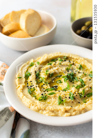 A bowl of creamy hummus topped with olive oil and herbs sits beside fresh bread and a bowl of olives. The light setting showcases a simple, inviting meal A bowl of creamy hummus topped with olive oil and herbs sits beside fresh bread and a bowl of olives. The light setting showcases a simple, inviting meal 131535930