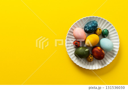 A variety of beautifully painted Easter eggs in different colors and patterns are placed on a delicate white plate. The vibrant hues stand out against the cheerful yellow backdrop A variety of beautifully painted Easter eggs in different colors and patterns are placed on a delicate white plate. The vibrant hues stand out against the cheerful yellow backdrop 131536030