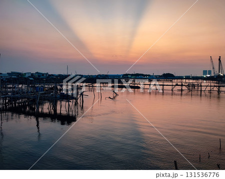 Beautiful sunset over a coastal village with dramatic sun rays, calm waters, and traditional fishing structures. Perfect for travel, nature, and scenic themes. 131536776