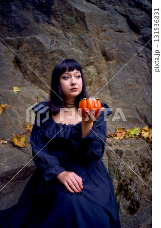 woman in a black long dress sits on black rocks on Halloween holding a pumpkin-shaped cup, a modern witch 131536831