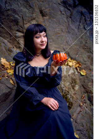 woman in a black long dress sits on black rocks on Halloween holding a pumpkin-shaped cup, a modern witch 131536832