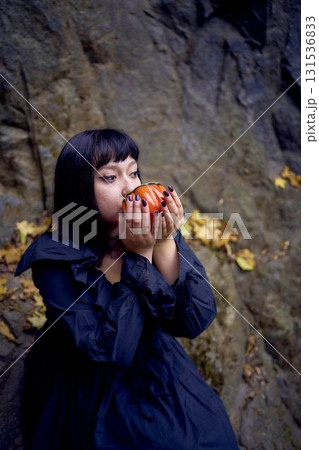 woman in a black long dress sits on black rocks on Halloween holding a pumpkin-shaped cup, a modern witch woman in a black long dress sits on black rocks on Halloween holding a pumpkin-shaped cup, a modern witch 131536833