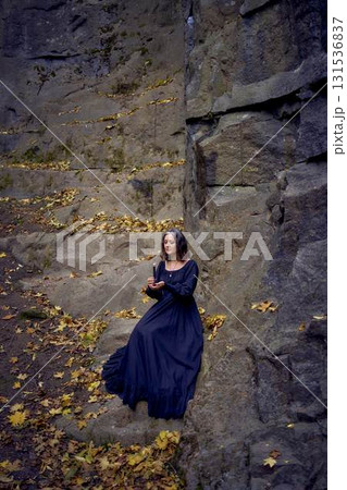 woman in a black long dress sits on black rocks on Halloween holding a burning candle, performs a witch ritual, modern witch 131536837