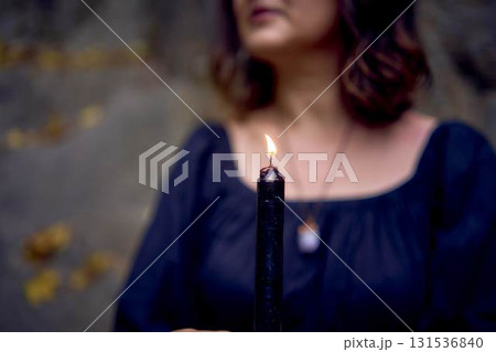 woman in a black long dress sits on black rocks on Halloween holding a burning candle, performs a witch ritual, modern witch woman in a black long dress sits on black rocks on Halloween holding a burning candle, performs a witch ritual, modern witch 131536840