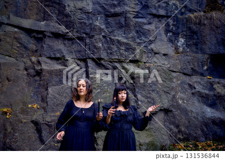 two women in the black long dresses near black rocks on Halloween holding a burning candle, performing a witch ritual, modern witch 131536844