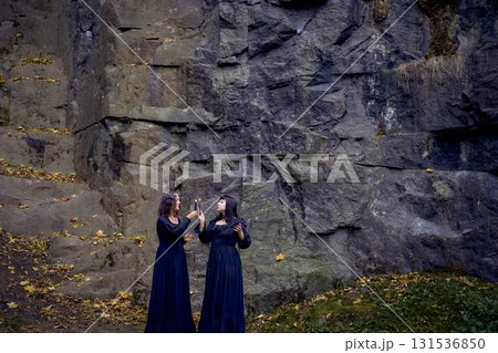 two women in the black long dresses near black rocks on Halloween holding a burning candle, performing a witch ritual, modern witch two women in the black long dresses near black rocks on Halloween holding a burning candle, performing a witch ritual, modern witch 131536850