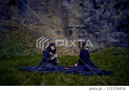 two women in black long dresses sitting on the black rocks on Halloween, reading tarot cards, performing a witch ritual, modern witch two women in black long dresses sitting on the black rocks on Halloween, reading tarot cards, performing a witch ritual, modern witch 131536856