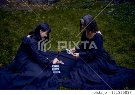 two women in black long dresses sitting on the black rocks on Halloween, reading tarot cards, performing a witch ritual, modern witch two women in black long dresses sitting on the black rocks on Halloween, reading tarot cards, performing a witch ritual, modern witch 131536857