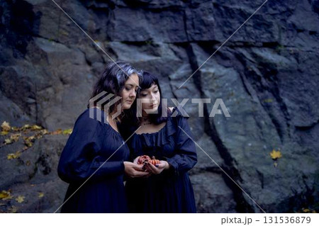 two women in the  black long dresses near black rocks on Halloween with candles in the form of pumpkins, performing a witch ritual, modern witch 131536879