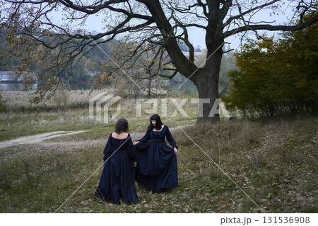 Two women in the long black dresses perform a historical reenactment of the Edwardian era dancing lovers on a hill near a majestic oak tree for Halloween. Two women in the long black dresses perform a historical reenactment of the Edwardian era dancing lovers on a hill near a majestic oak tree for Halloween. 131536908