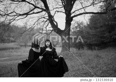 Two women in the long black dresses perform a historical reenactment of the Edwardian era dancing lovers on a hill near a majestic oak tree for Halloween. 131536910