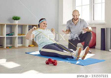 Friendly nurse helping fat woman doing stretching exercises with rubber band sitting on the floor Friendly nurse helping fat woman doing stretching exercises with rubber band sitting on the floor 131537221