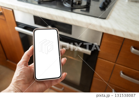 Hand holding smartphone with white blank screen near modern kitchen oven. Woman operating through mobile application. Smart home. Close up 131538087