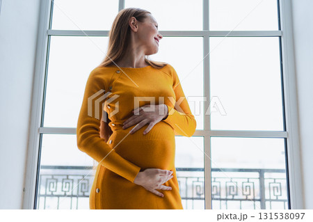 Pregnancy motherhood people expectation future. Pregnant woman with big belly standing near window at home. Girl hugging her tummy enjoying pregnancy. Maternity tenderness parenthood new life concept 131538097