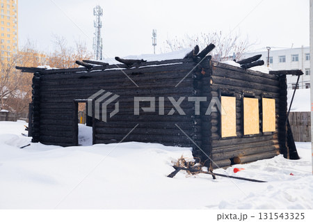 Burnt abandoned wooden house with boarded up windows and snow. Nearby, buildings contrast sharply with the rustic cabin, reflecting urban development. Building to be demolished Burnt abandoned wooden house with boarded up windows and snow. Nearby, buildings contrast sharply with the rustic cabin, reflecting urban development. Building to be demolished 131543325