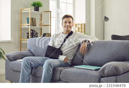 Happy, smiling man with orthopedic sling on injured arm sitting on couch at home Happy, smiling man with orthopedic sling on injured arm sitting on couch at home 131546470