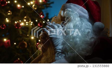Santa Claus Seated On The Sofa And Drinks Tea Next To The Illuminated Tree 131555660