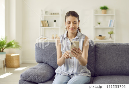 Young woman sitting on couch at home and browsing social media on her cell phone 131555793