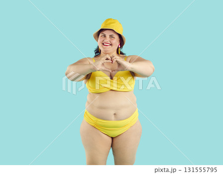 Funny young fat woman inyellow bikini doing heart symbol shape with hands on blue background. Funny young fat woman inyellow bikini doing heart symbol shape with hands on blue background. 131555795