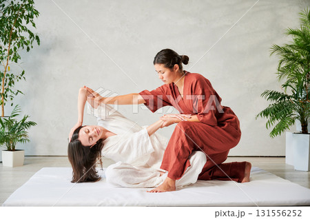 Masseuse making thai yoga massage. Woman therapist doing traditional massage treatment, stretching female patient's body. Client sitting on mat, enjoying hand massage in calm atmosphere. 131556252